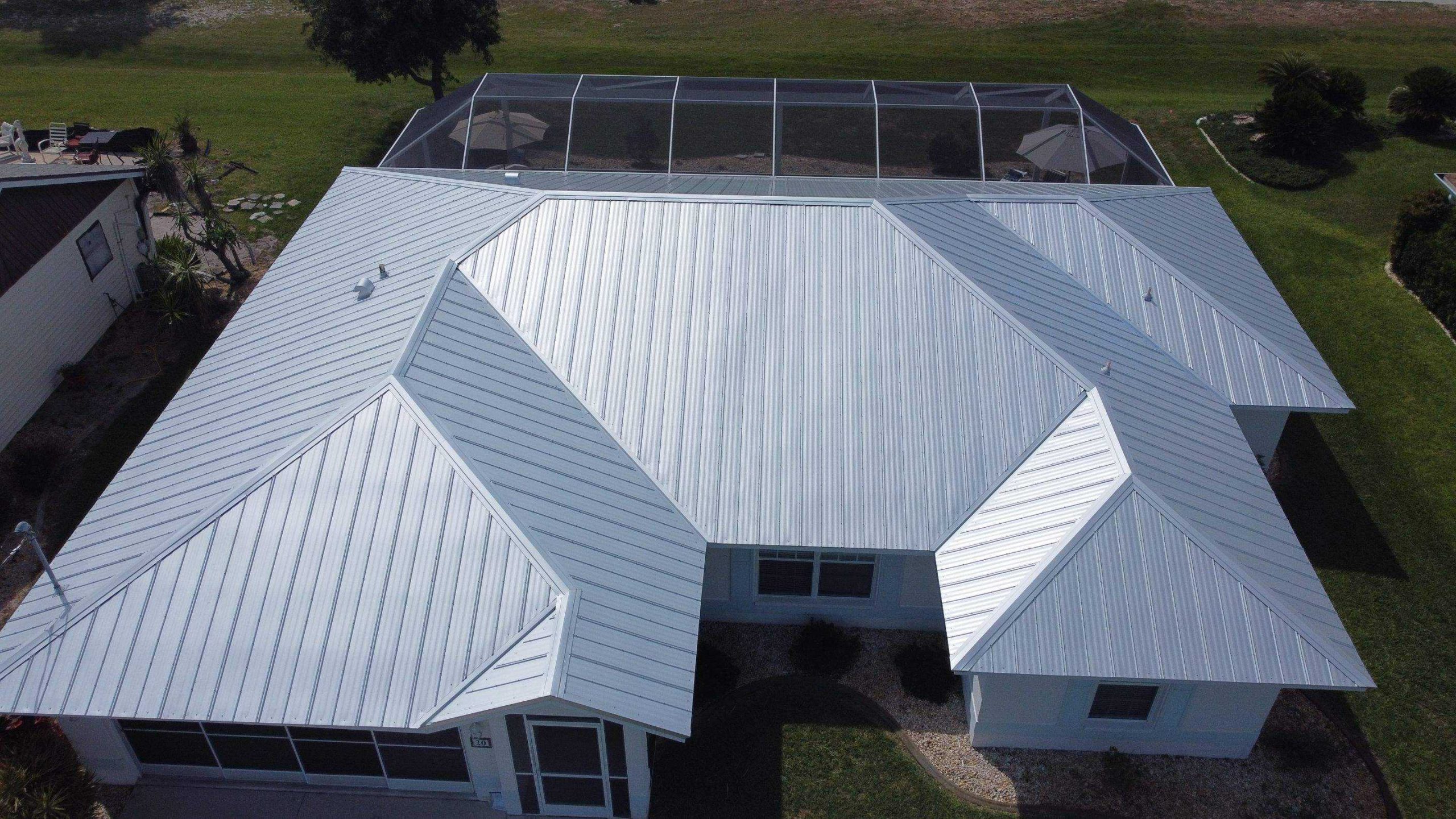 Aerial view of a white, metal-roofed house with multiple gables. The backyard features a screened-in patio area. The property, perfect for a roofer in Sarasota County, is surrounded by green grass and a neighboring house is partially visible. The sky appears clear, providing ample sunlight on the roof.