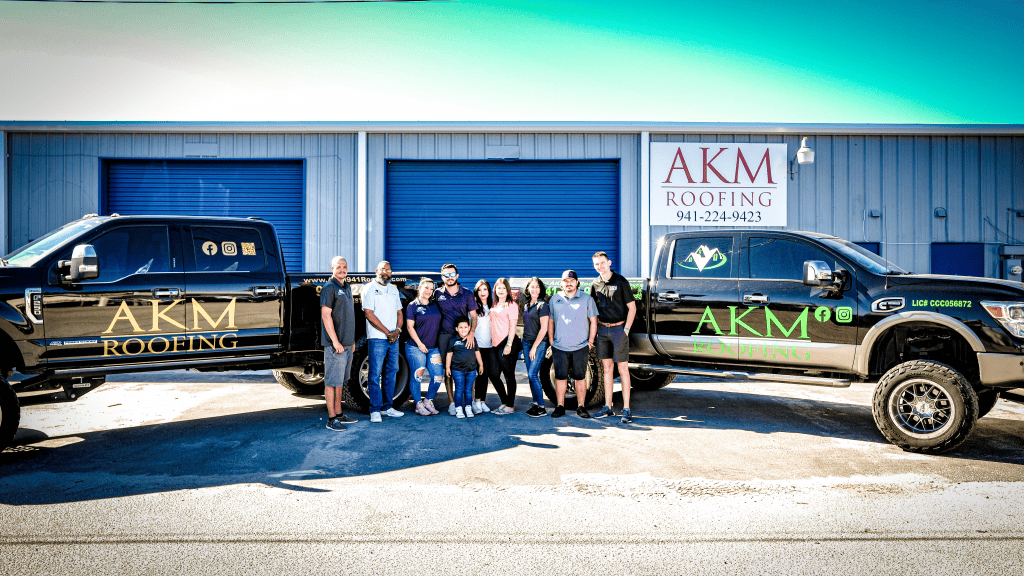 A group of nine people stands in front of a building with two black trucks on either side, all featuring "AKM Roofing" logos. The smiling team represents the top roofing contractor Sarasota County trusts. The building has two large blue doors and a sign with a phone number.
