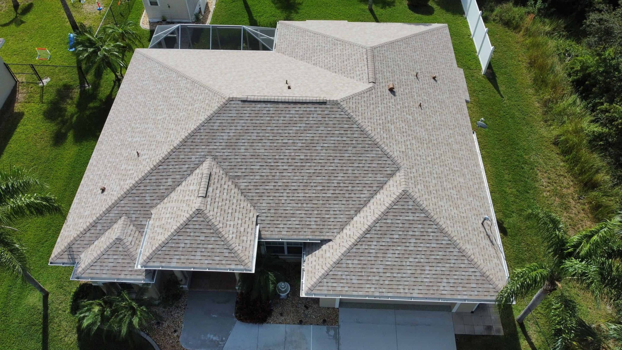 Aerial view of a residential house with a gabled roof design and light-colored shingles. The property, possibly serviced by a Roofing Contractor in Sarasota County, is surrounded by well-maintained green grass and a few palm trees. A driveway leads to the house entrance.