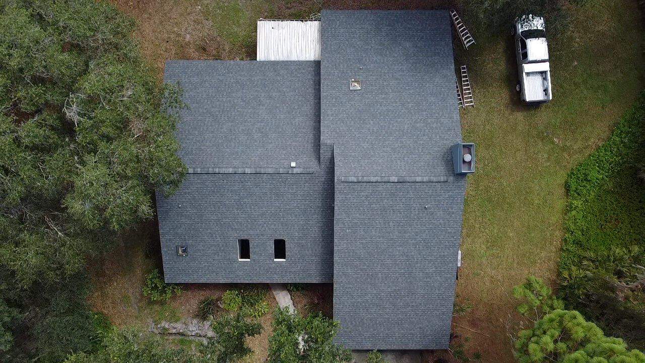 Aerial view of a house with a dark gray roof surrounded by trees and greenery. There is a white pickup truck parked on the lawn, along with several ladders leaning against the house, likely for roof repair in Sarasota County. The backyard features a small, light-colored deck.