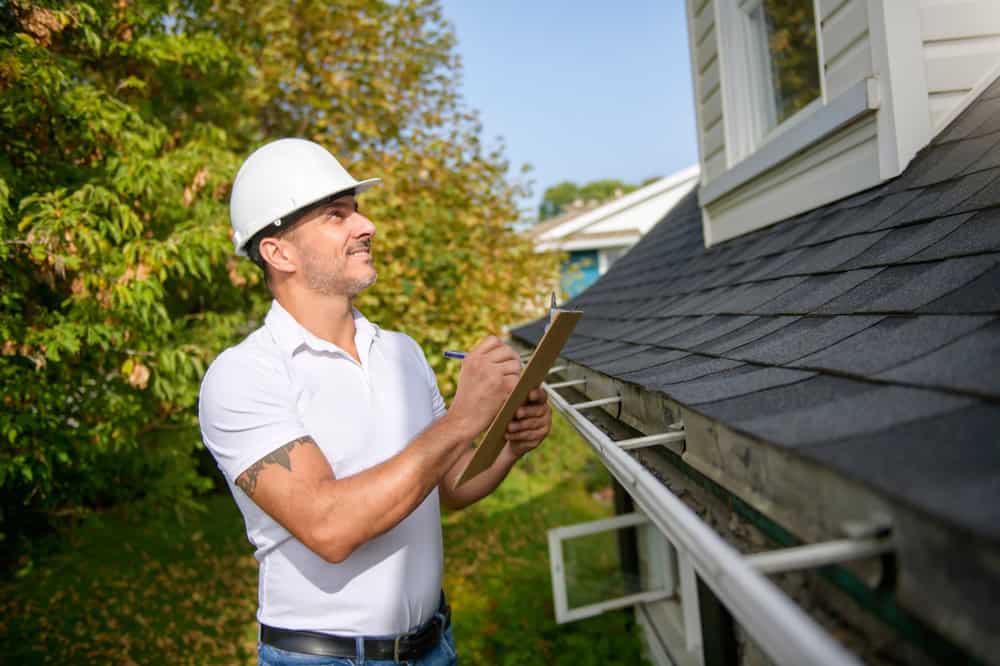 A man wearing a white hard hat and a white polo shirt is inspecting a house, conducting a roof inspection in Sarasota County. He is holding a clipboard and pen, looking up at the roof. The house is white with gray shingles. Trees can be seen in the background, indicating a residential area.
