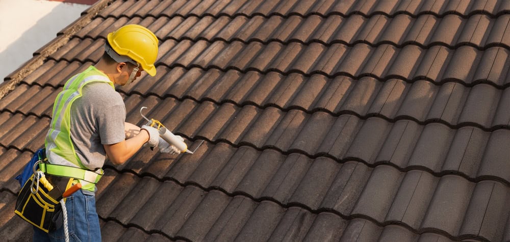 A construction worker wearing a yellow hard hat and safety harness applies sealant to the tiles of a dark brown roof. He's using a caulking gun and standing at the roof's edge on a sunny day.